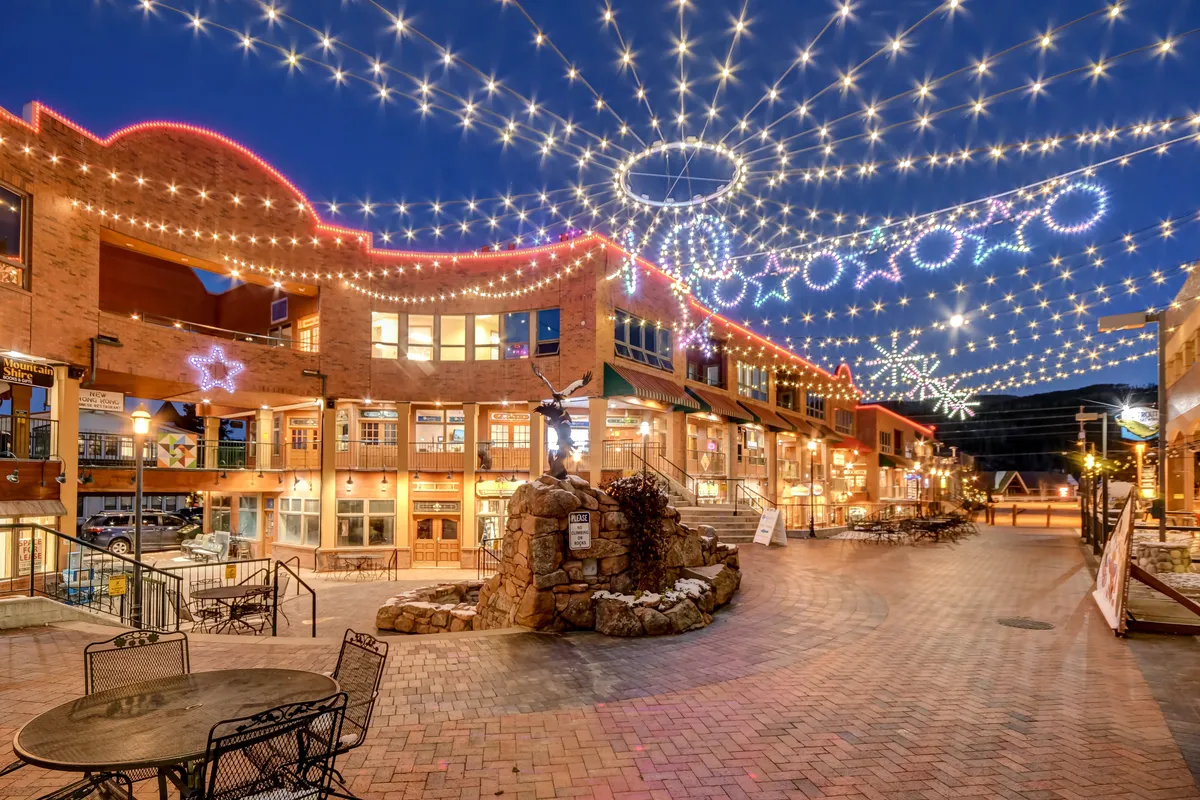 A lively downtown Winter Park scene at dusk, with twinkling decorative lights strung above a central fountain, surrounded by shopfronts bustling with diners, shoppers, and friends enjoying the festive evening. The overall mood is joyful and social, capturing the spirit of local nightlife and community events.