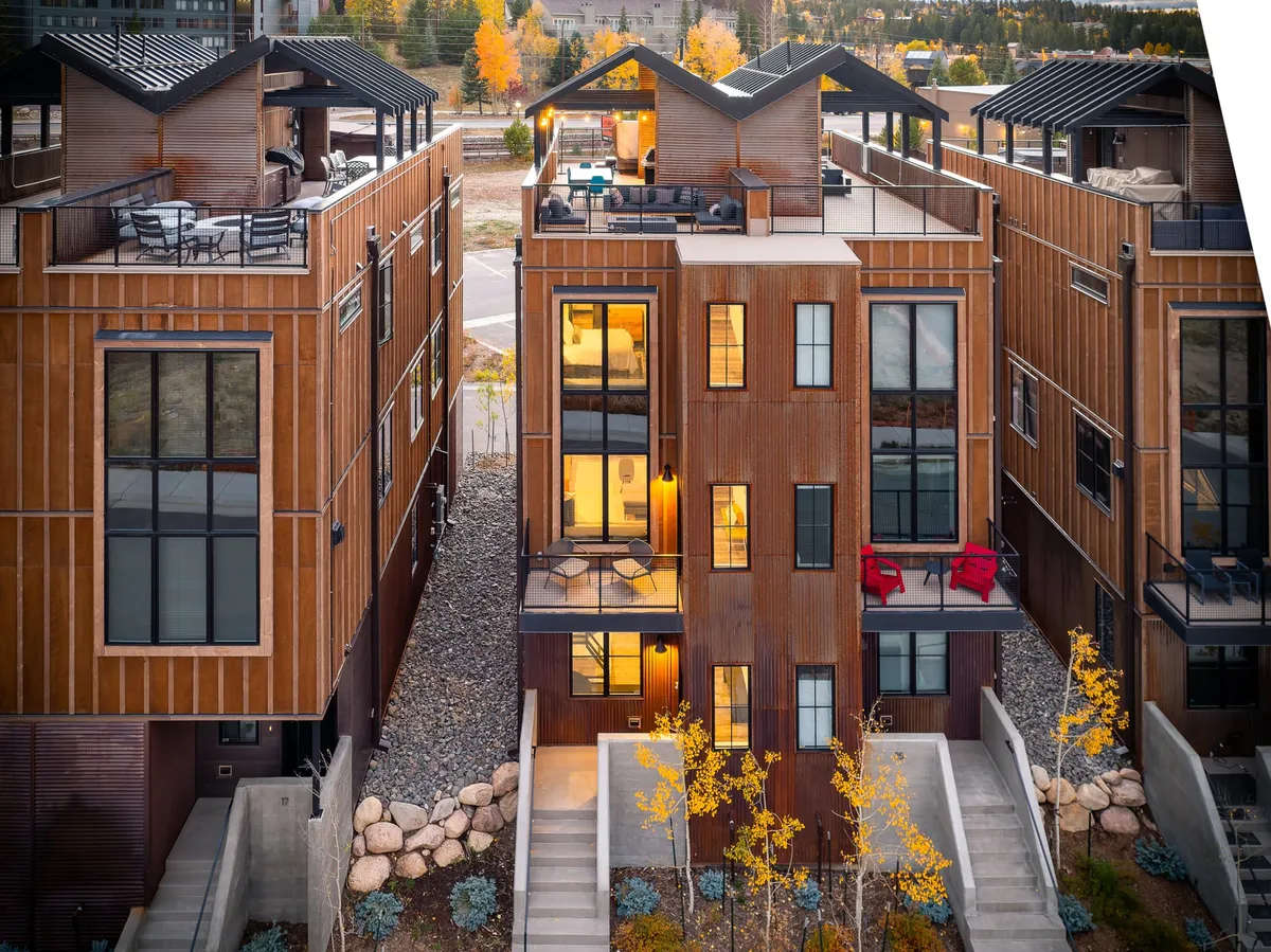 The image showcases the contemporary exterior of a luxury loft in Winter Park, Colorado, featuring glowing windows that suggest warmth and comfort inside. The home displays a striking combination of wood and metal elements, with inviting rooftop terraces perfect for socializing or relaxing under the stars. Evening lighting sets a cozy, welcoming mood, while the surrounding mountain landscape hints at adventure and outdoor activities.
