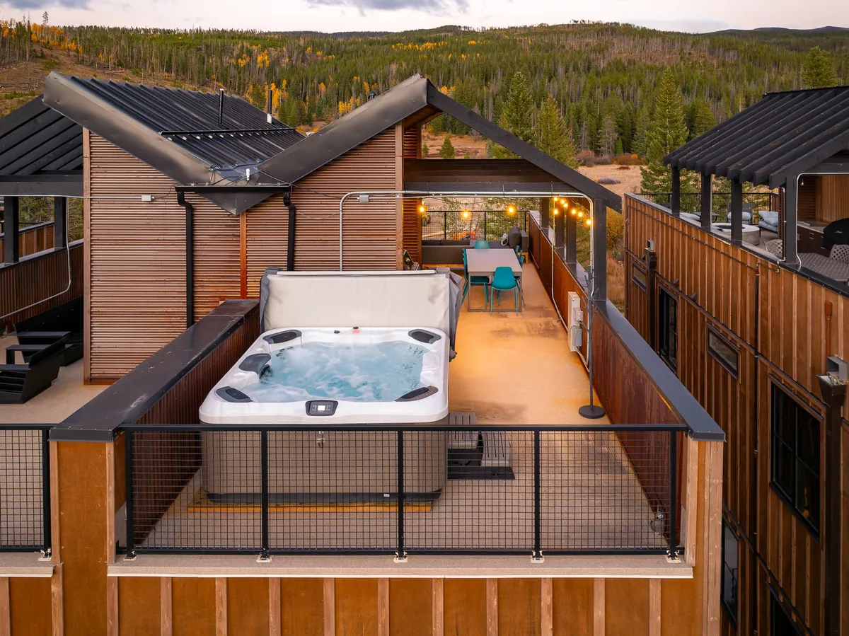 An inviting outdoor scene featuring a private hot tub and dining area surrounded by trees, with string lights creating a cozy atmosphere. The tranquil setting is perfect for relaxing after a day of adventure, with views of nature all around.
