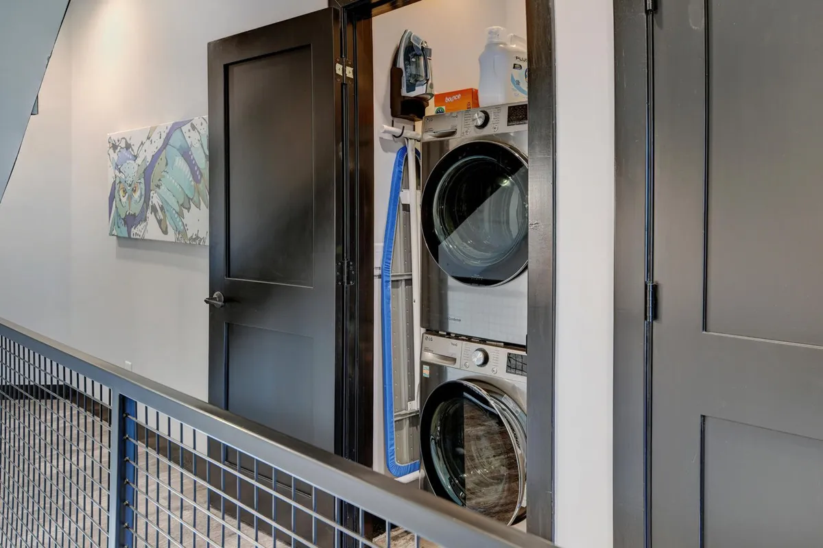 A neat laundry area featuring a stacked washer and dryer, iron and ironing board, and colorful artwork, providing a practical space to freshen up clothes during longer stays.