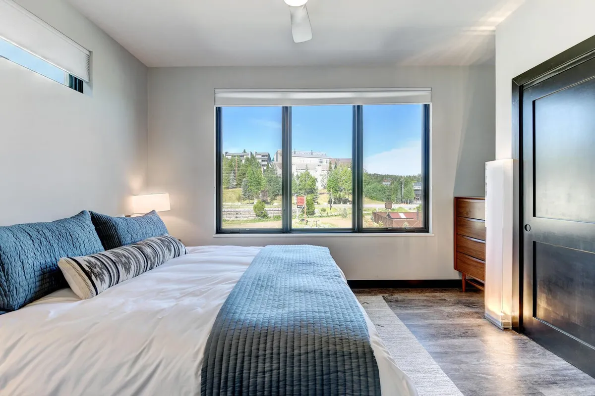 A serene primary bedroom with a king-sized bed dressed in blue bedding, large windows offering a scenic view, and a modern dresser. Natural light fills the space, creating a tranquil atmosphere for relaxation and rest.