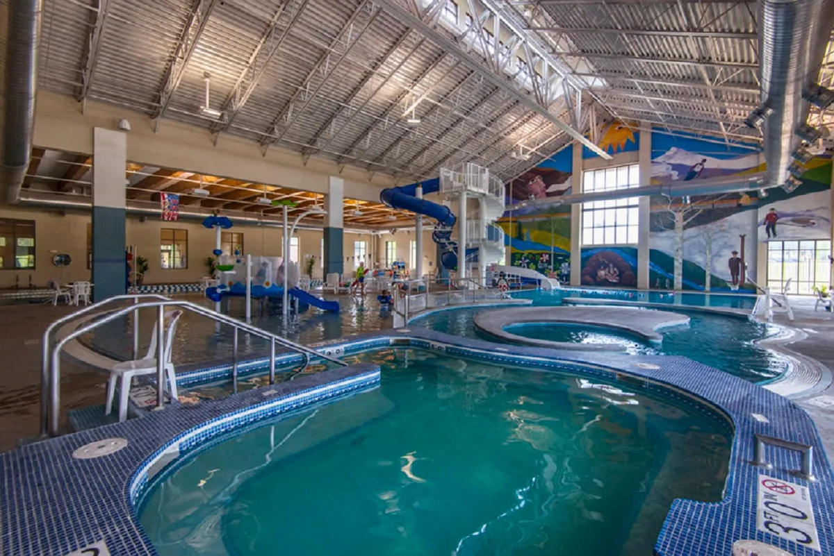 A vibrant indoor pool area at the Grand Park Rec Center, with water slides, children playing, and families gathered around. The colorful setting radiates joy and recreation.