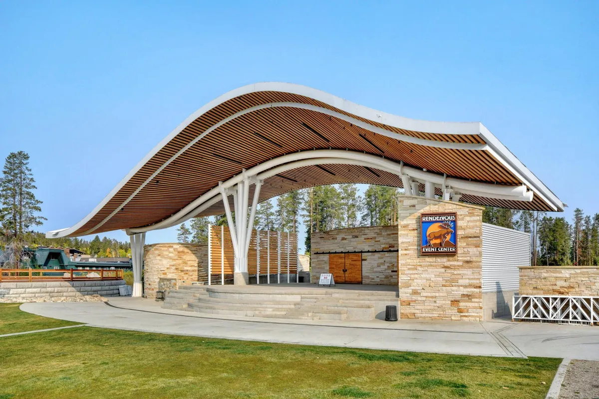 This exterior image depicts a striking curved roof structure with a stone facade and an open stage area, set up for outdoor performances and community events. The scene exudes a celebratory mood, suggesting lively gatherings and local festivities.