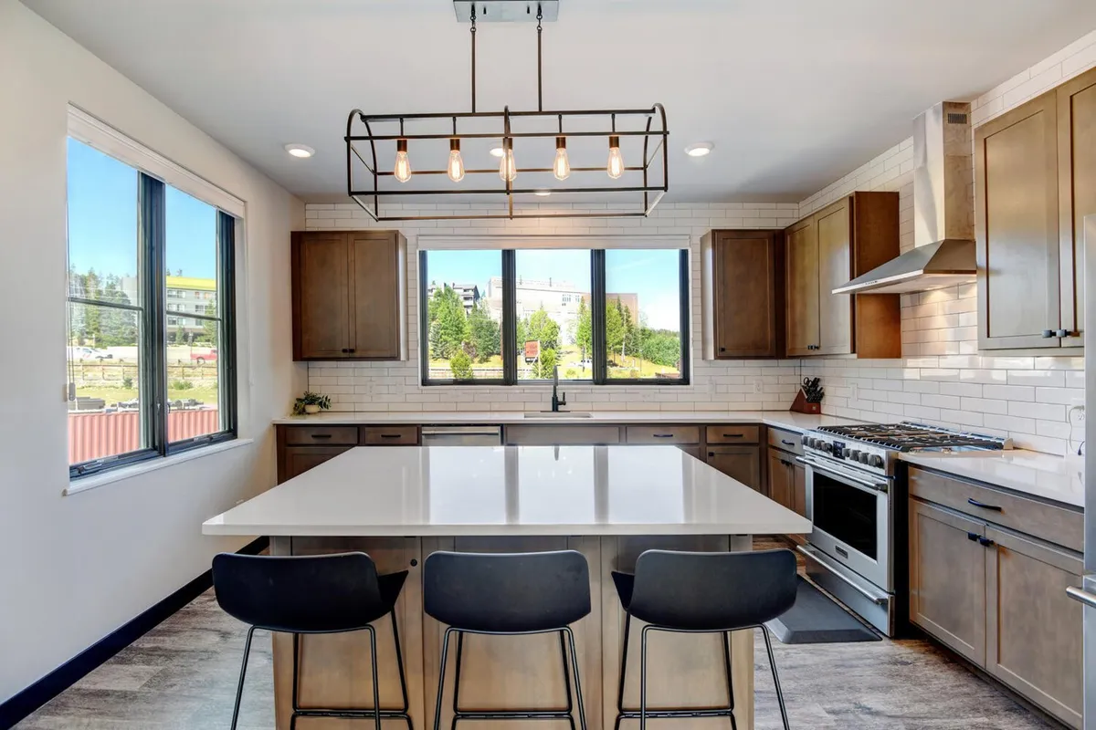 A spacious, light-filled kitchen featuring a large island countertop with bar seating, gleaming high-end appliances, modern lighting fixtures, and plenty of windows letting in natural light. The layout invites interaction and culinary creativity, creating an ideal space for both cooking and gathering.