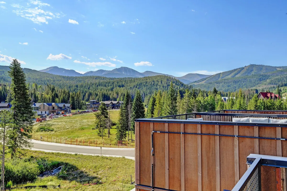 A panoramic view of a majestic mountain range bordered by thick forests, with a modern building and winding roadway nestled in the landscape. The expansive view emphasizes the scale and beauty of the Rockies and hints at opportunities for hiking, biking, and nature exploration.