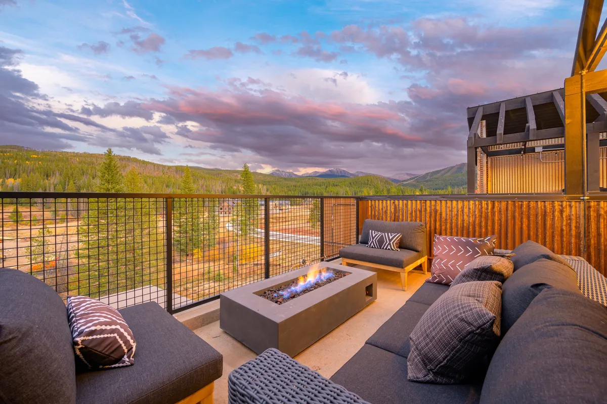 A modern fire pit is surrounded by plush outdoor seating on a rooftop patio, with stunning mountain views stretching into the distance. The inviting setup hints at cozy gatherings, perfect for stories and s’mores as night falls.